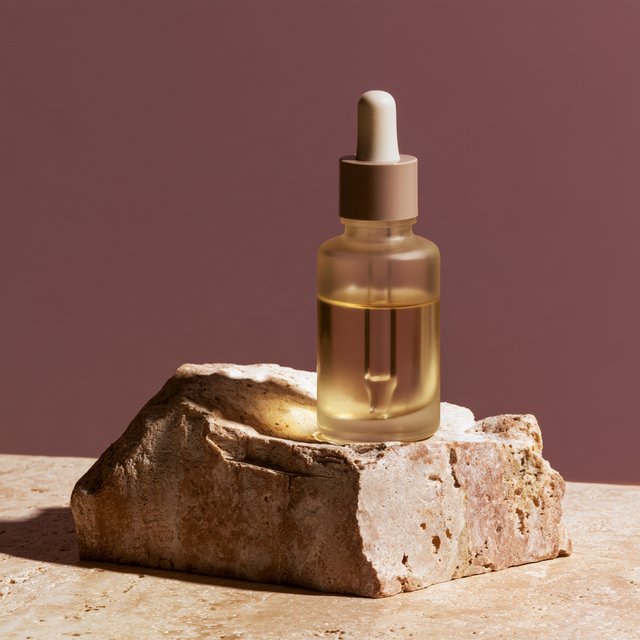 Under a broad diffused key and cool edge light, an unbranded frosted-glass serum bottle displays clear golden oil against a muted plum background, resting on honed pale travertine with its blank surface free of lettering. — Midjourney V8.2
