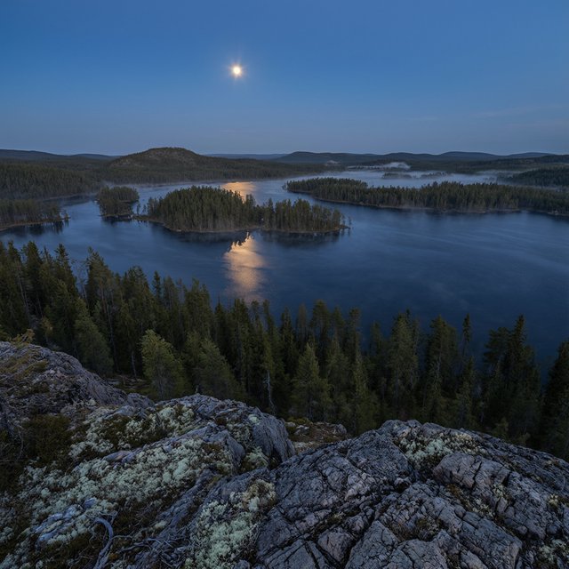 An expansive boreal lake beneath a clear, cold midnight sky, pale moonlight catching thin mist above dark water; spruce-covered islands recede toward distant ridges, while lichen and cracked granite sharpen the near shoreline. — Qwen Image 3.0 Pro