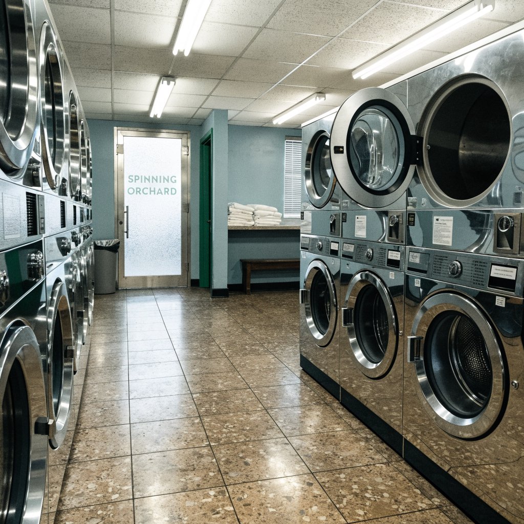 Inside a quiet neighborhood laundromat, chrome machines and folded linen glow beneath cool ceiling lights; "SPINNING ORCHARD" is printed in pale mint letters on the frosted glass entry door. — Recraft v4