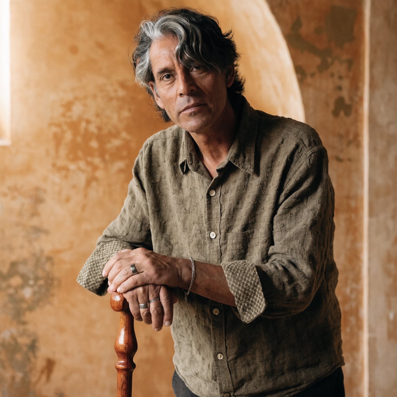 Portrait photograph of an adult man in his 40s with silver-streaked hair, wearing a textured olive linen shirt and rolled sleeves, hands resting on a wooden cane, relaxed expression, ochre plaster backdrop, soft window light from the side. — Recraft v4 Pro