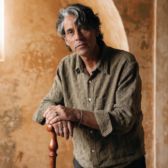 Portrait photograph of an adult man in his 40s with silver-streaked hair, wearing a textured olive linen shirt and rolled sleeves, hands resting on a wooden cane, relaxed expression, ochre plaster backdrop, soft window light from the side. — Recraft v4 Pro