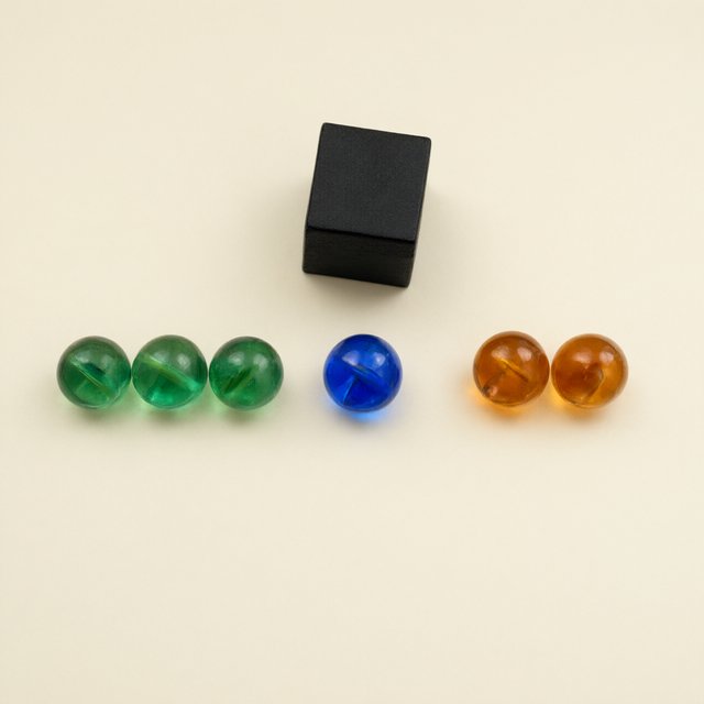 Exactly six glass marbles sit on a cream tabletop: three green marbles in a row on the left, two amber marbles in a row on the right, and one blue marble centered between the rows. A black cube sits above the blue marble, nothing else. — Qwen Image 3.0
