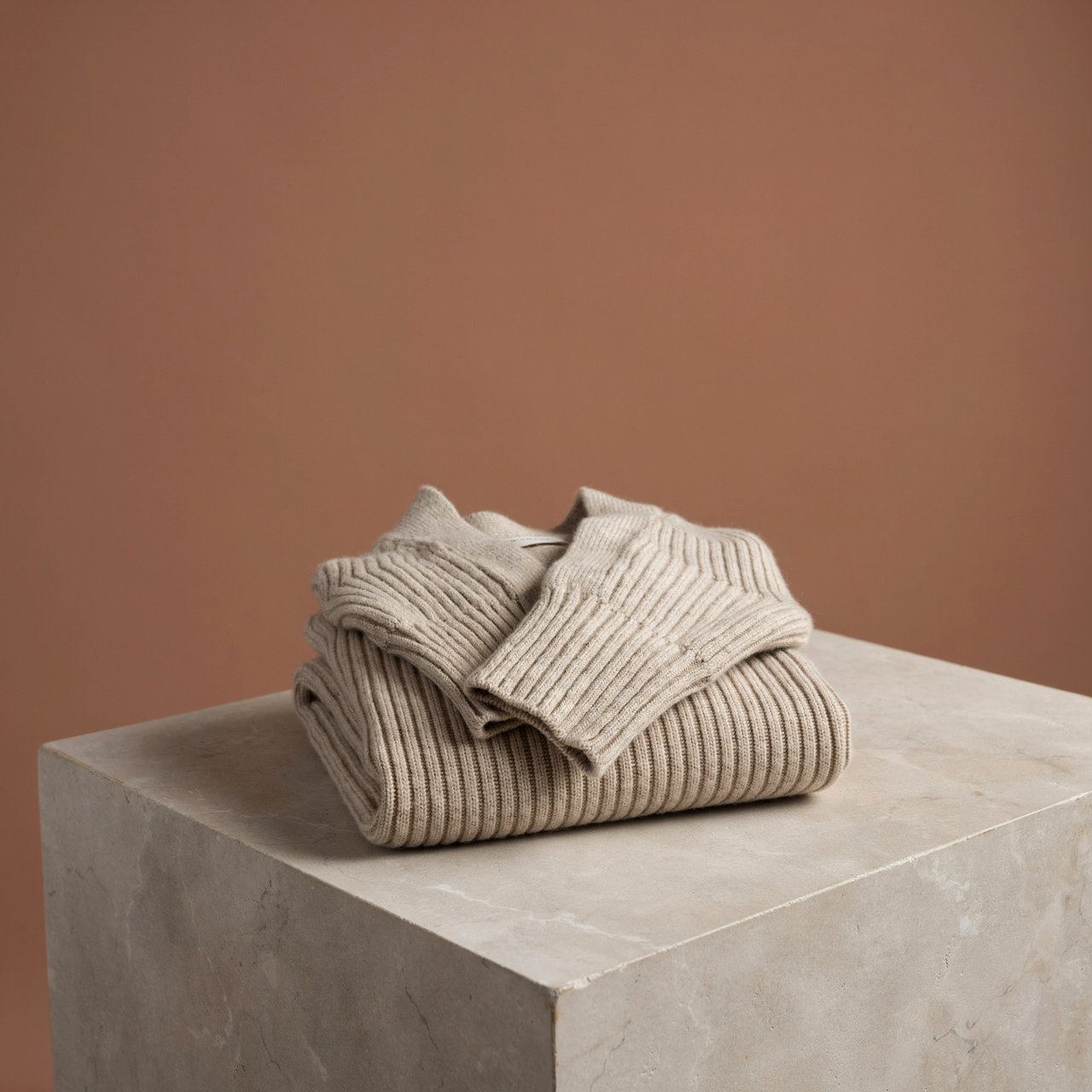 A folded unbranded merino cardigan rests on a honed limestone plinth, its chunky ribbed wool surface softly catching broad overhead diffusion and warm side bounce against a muted clay backdrop, with no tags, lettering, or graphics. — Ideogram v4