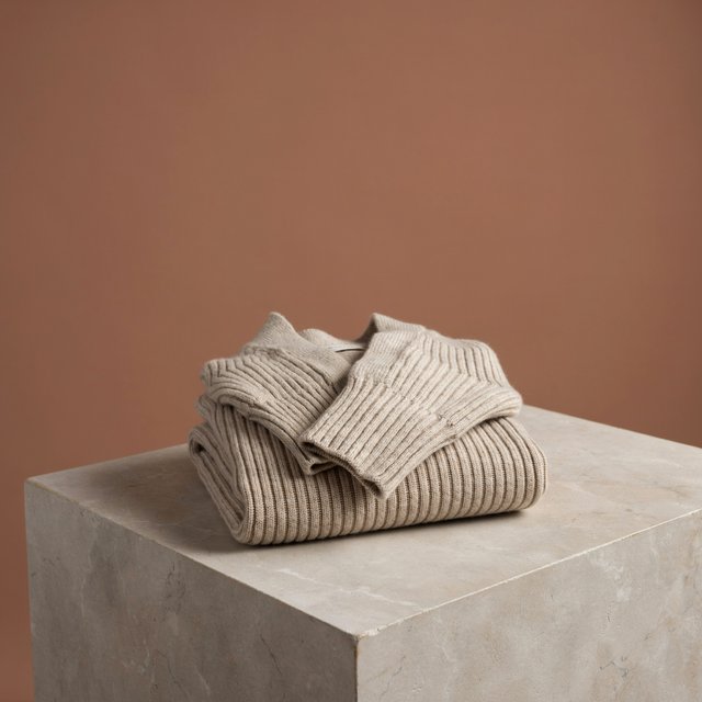 A folded unbranded merino cardigan rests on a honed limestone plinth, its chunky ribbed wool surface softly catching broad overhead diffusion and warm side bounce against a muted clay backdrop, with no tags, lettering, or graphics. — Ideogram v4