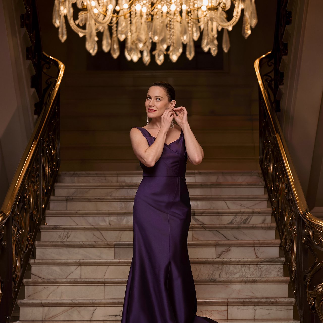 At the landing of a marble staircase, an adult woman in a plum evening gown pauses to fasten an earring, caught in warm chandelier light; polished brass rails and soft shadows lend the scene the poise of a fashion magazine. — Kling v3