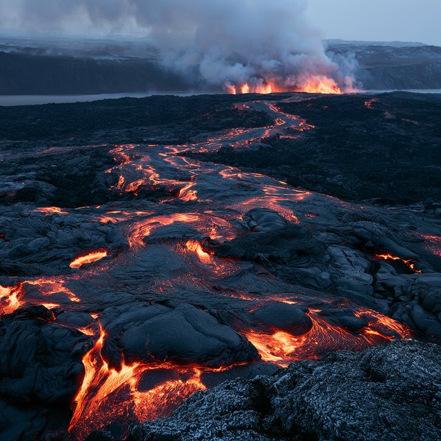 Fresh lava threads a black cinder plain on a volcanic island at dusk beneath ash-laden wind; blistered crust and brittle pumice in the foreground give way to glowing fissures fading into smoke. — Midjourney V8.2