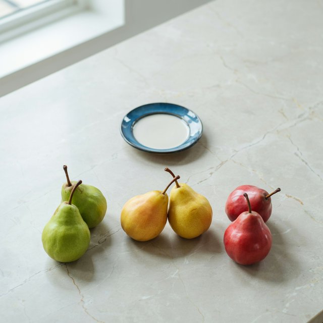 Exactly six ripe pears sit on a pale stone counter: two green pears on the left, two yellow pears in the center, and two red pears on the right. A small blue plate sits above the center pair, nothing else. — Ideogram v4