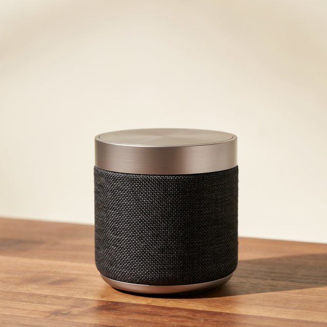 Commercial product photograph of an unbranded wireless speaker made from textured black fabric and satin aluminum, placed on a walnut tabletop against a warm cream background, illuminated by a large softbox and subtle backlight. — Ideogram v4