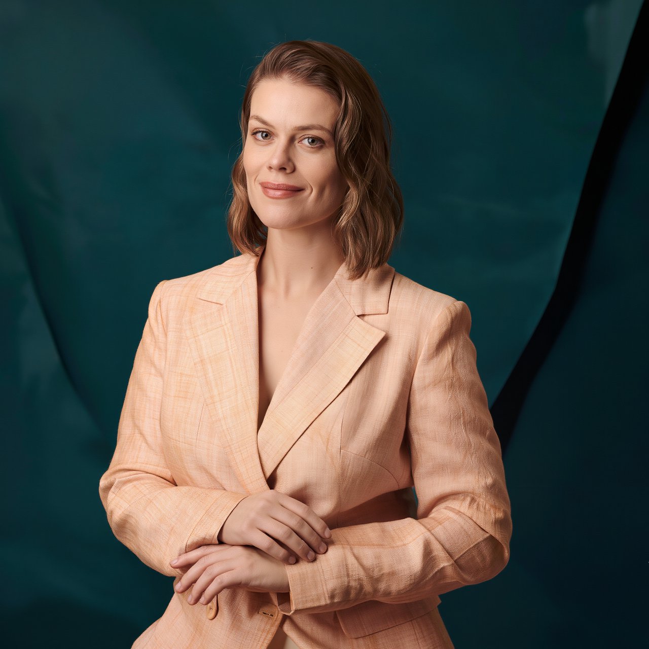 Portrait photograph of an adult woman in her early 30s wearing a pale apricot linen blazer, her hands gently smoothing its cuff. She looks quietly delighted against a deep teal paper backdrop, illuminated by soft, angled studio light. — Kling v3