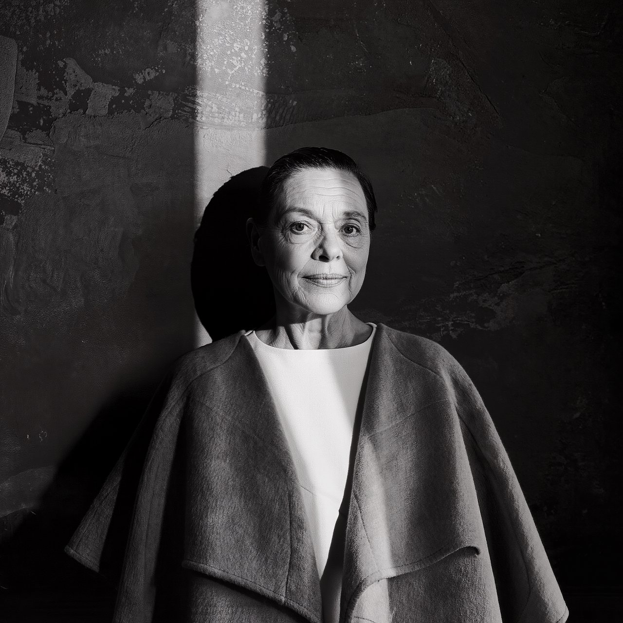 Under a narrow window beam, an adult woman in her late 50s wears a sculpted wool cape over a cotton blouse; her wry expression rests against a charcoal plaster backdrop in a black-and-white portrait, shaped by soft angled light. — Kling v3