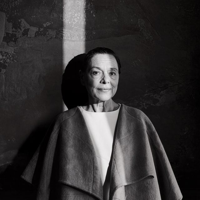 Under a narrow window beam, an adult woman in her late 50s wears a sculpted wool cape over a cotton blouse; her wry expression rests against a charcoal plaster backdrop in a black-and-white portrait, shaped by soft angled light. — Kling v3