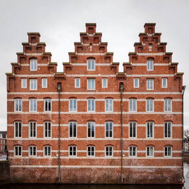 Exterior of a Dutch Renaissance canal-side archive, with warm red brick, pale stone bands, stepped gables, and tall sash windows; soft overcast light reveals the masonry, viewed from the opposite quay, with convincing structure and precise architectural lines. — Kling v3