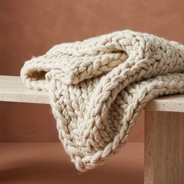 Soft folds of an unbranded chunky-knit wool throw rest on a pale travertine bench; its looped surface catches broad diffused key light and gentle side fill against a muted terracotta studio background, forming a clean commercial product image with no lettering. — Model 15