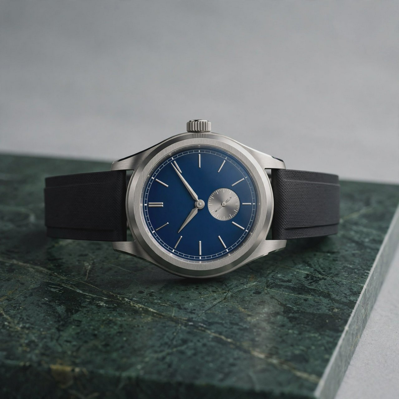 Commercial product photograph of an unbranded mechanical wristwatch with a brushed titanium case and deep blue enamel dial, resting on dark green marble against a soft gray background, illuminated by a large diffused softbox and narrow edge light. — Muse Image 1.0