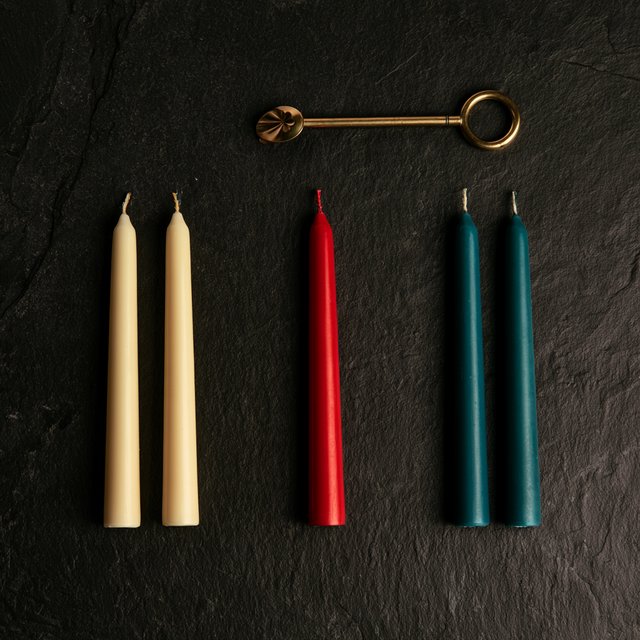 Exactly five candles stand on a dark slate tabletop: two ivory candles on the left, one red candle in the center, and two teal candles on the right. A brass snuffer sits above the red candle, nothing else. — Midjourney V8.2