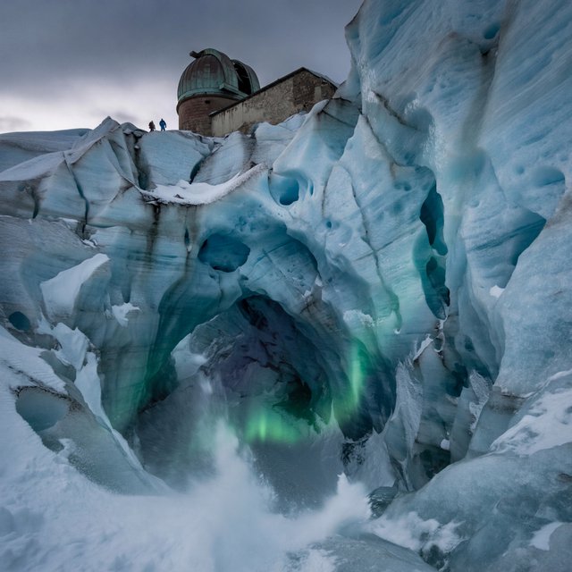 An abandoned observatory carved into a glacier, its bronze domes protruding from blue ice, aurora light filtering through crevasses, tiny adult explorers on a distant ridge emphasizing immense scale, drifting snow and deep polar shadows. — ImagineArt 2