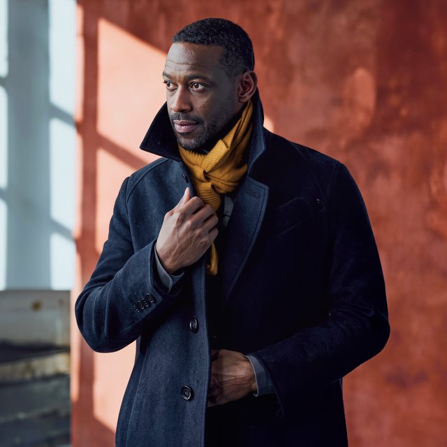Editorial portrait of an adult Black man in his 30s wearing a deep navy wool overcoat and mustard scarf, fingers adjusting the scarf, thoughtful expression, muted terracotta studio backdrop, crisp cool window light. — Kling v3