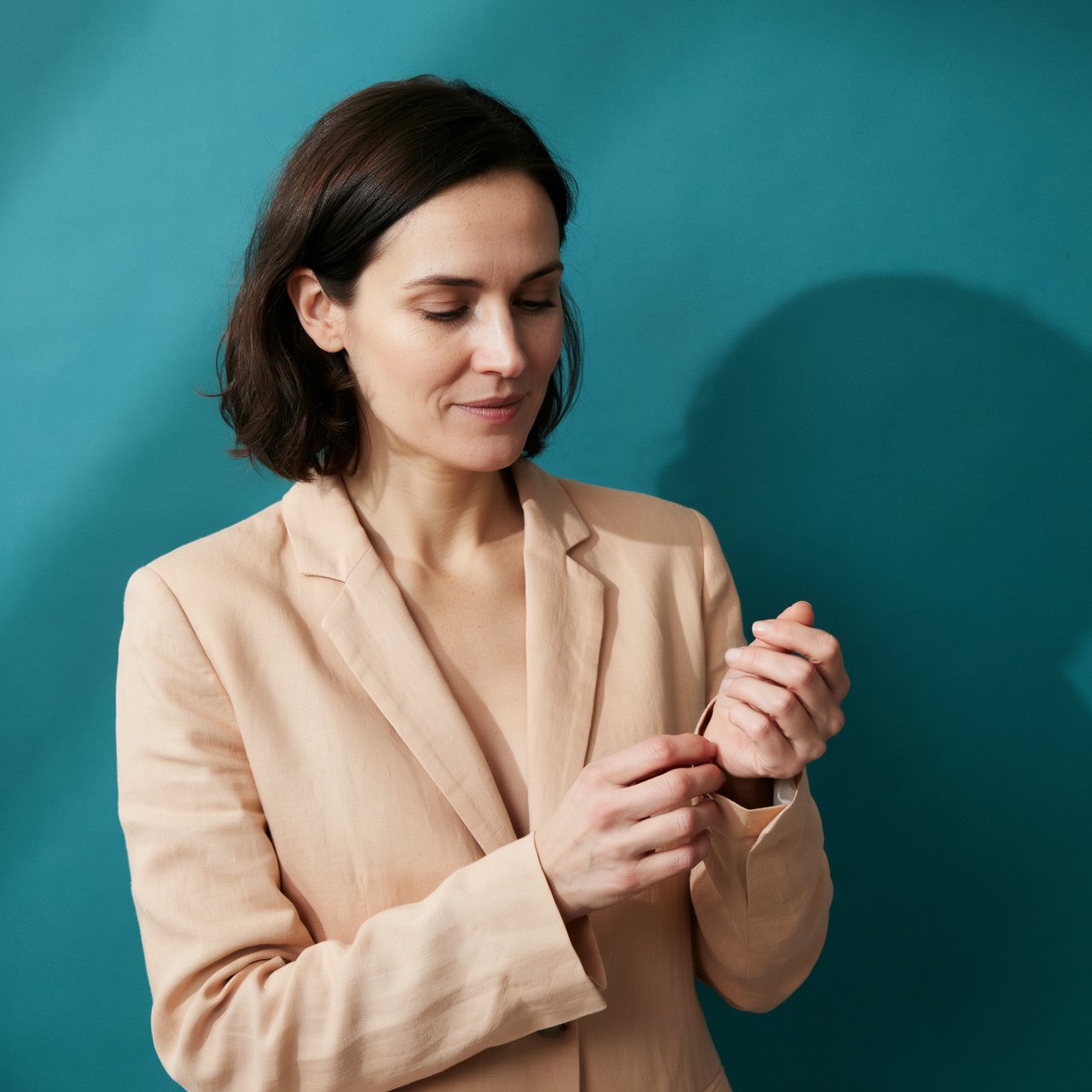 Portrait photograph of an adult woman in her early 30s wearing a pale apricot linen blazer, her hands gently smoothing its cuff. She looks quietly delighted against a deep teal paper backdrop, illuminated by soft, angled studio light. — Ideogram v4