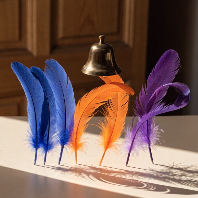 Exactly seven feathers lie on a cream tabletop: three blue on the left, two orange in the center, and two purple on the right. A brass bell sits above the orange pair, nothing else. — Recraft v4 Pro