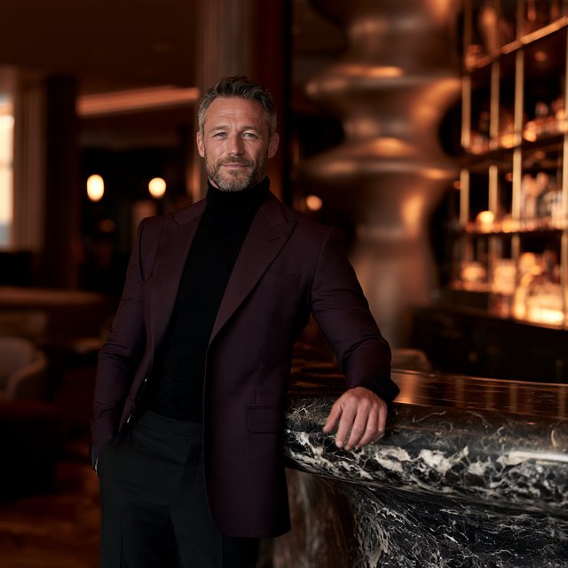 Editorial glamour photograph of an adult man in a midnight-plum dinner jacket and black trousers, standing naturally beside a sculptural marble bar in a quiet hotel lounge, flattering amber light, easy smile, polished editorial elegance. — Midjourney V8.2