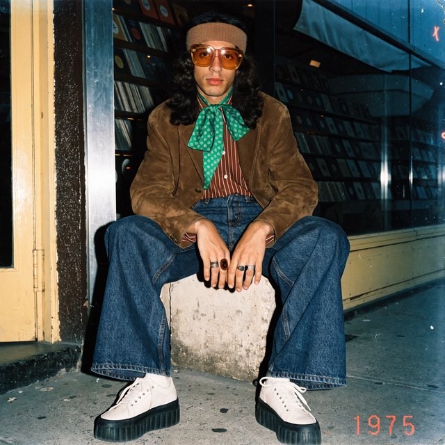 Create an image of a man in the quintessential style of 1975. Style his hair, clothing, accessories, and surroundings to reflect the distinctive look of the mid-1970s—featuring bold silhouettes, eye-catching accessories, layered details, and characteristic colors and textures. Give the image the feel of an authentic 1975 photograph, complete with analog film grain, faded colors, direct flash lighting, and a slight soft-focus effect. Add a period-accurate, red-orange date stamp in a 1970s style in one of the bottom corners. No recognizable posters. No modern objects or text. — Recraft v4 Pro