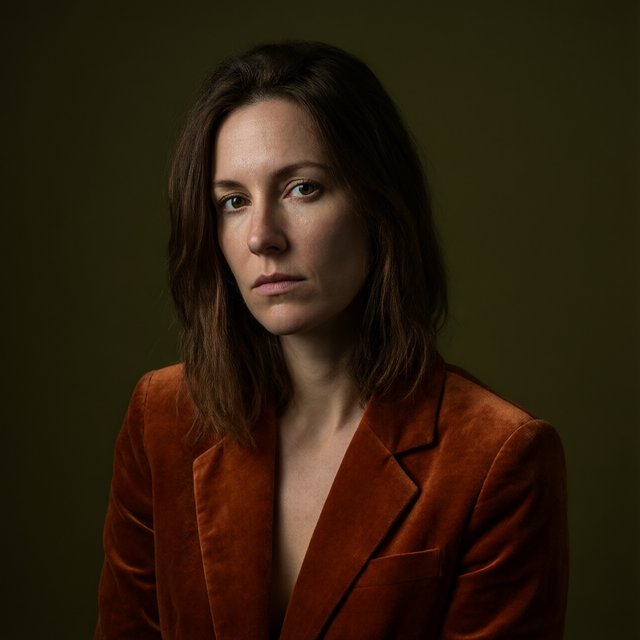 Her guarded gaze catches a solitary beam in a low-key fashion portrait: an adult woman aged 27–35 wears a rust-colored velvet blazer, the rest of her face falling into shadow against a deep olive backdrop. — Qwen Image 3.0