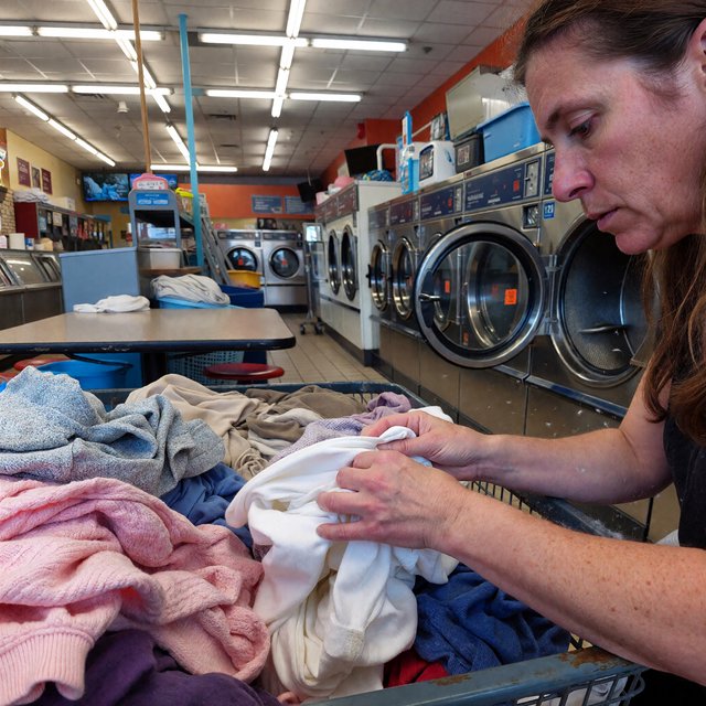 Candid documentary photograph of an adult sorting laundry in a neighborhood laundromat, cool fluorescent light, 35mm lens with moderate depth of field, lint specks floating near a tumbling dryer door. — ImagineArt 1.5 Pro