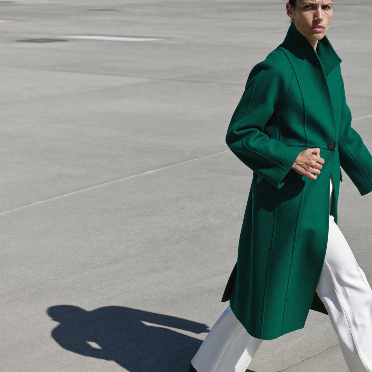 Full-length fashion editorial: model in a structured emerald wool coat and white trousers walking across a wide empty plaza, harsh midday sun, long shadow. — ImagineArt 2