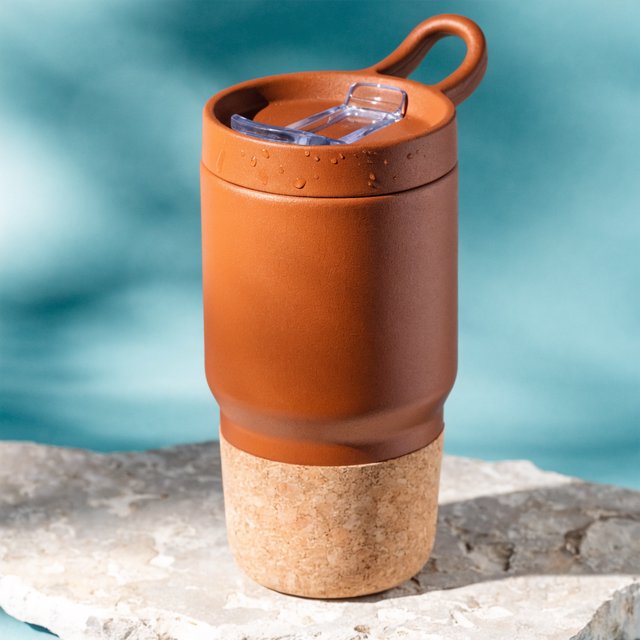 Commercial product photograph of an unbranded insulated travel mug made from matte terracotta powder-coated steel with a cork grip, standing on a pale limestone slab against a soft teal background, lit by broad diffused window light and a gentle edge light. — Recraft v4