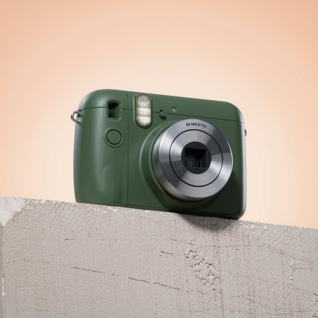 Commercial product photograph of an unbranded compact instant camera in matte forest-green polymer with brushed steel details, resting on a pale concrete surface against a warm peach background, illuminated by broad diffused studio light and a soft rim light. — Kling v3 Omni