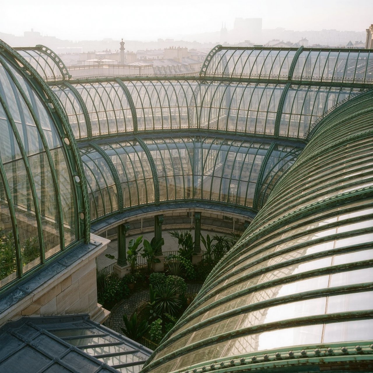 From a nearby rooftop, an Art Nouveau conservatory curves around a planted courtyard, its green copper ribs, clear glass, and pale stone base catching hazy morning light; orderly rooflines and believable supports show from above. — Kling v3 Omni