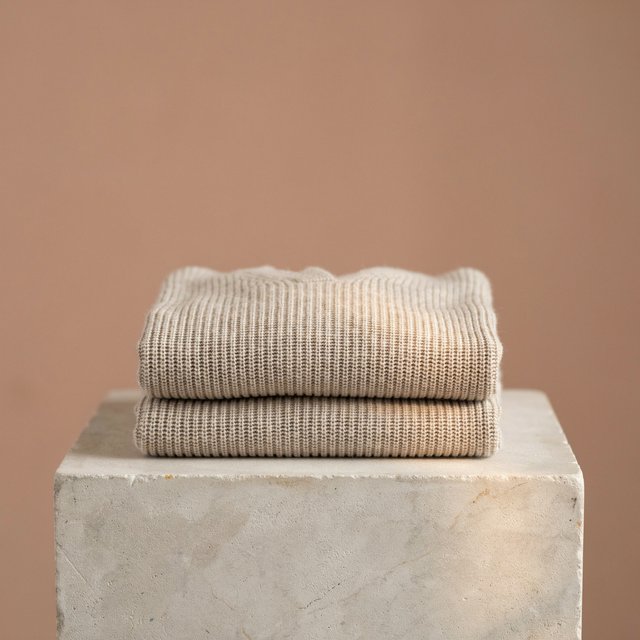 A folded unbranded merino cardigan rests on a honed limestone plinth, its chunky ribbed wool surface softly catching broad overhead diffusion and warm side bounce against a muted clay backdrop, with no tags, lettering, or graphics. — Ideogram v4.5
