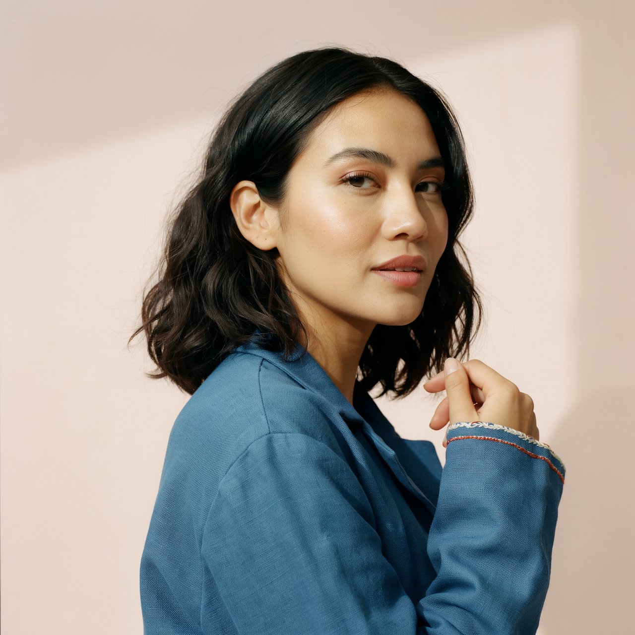Fashion portrait of an adult Latina woman in her late 20s wearing a handwoven cobalt-blue cotton jacket, one hand touching its embroidered cuff, curious half-smile, against a pale peach studio backdrop, soft diffused window light shaping her face. — Ideogram v4