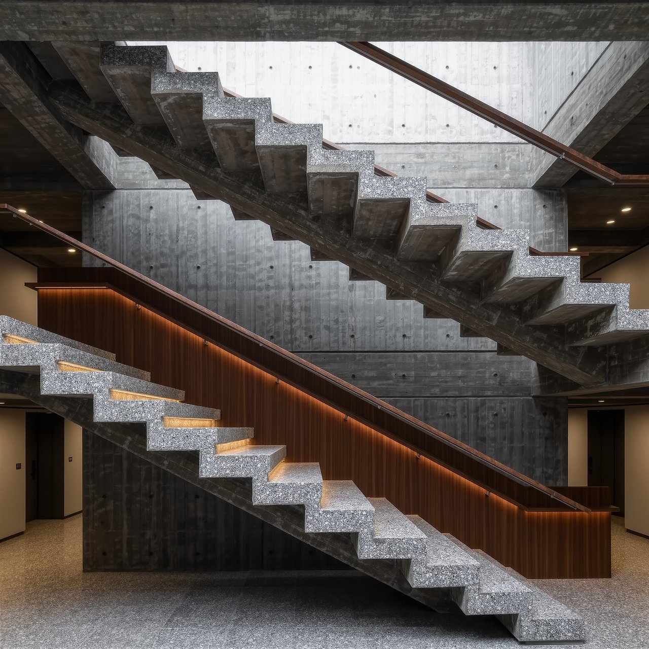 At the heart of a restrained Brutalist museum, a folded concrete stair rises beside smoked-oak handrails and terrazzo landings; skylight washes the treads, seen from the lower landing, with aligned flights and believable supports. — Kling v3 Omni