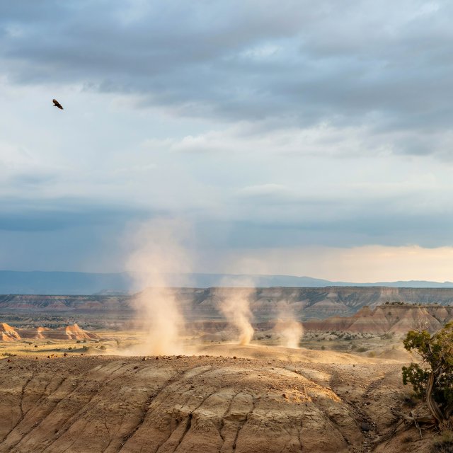 Wind-driven dust devils spiral across ochre badlands beneath a storm-clearing late-afternoon sky; pebble-studded clay ridges and tiny erosion grooves sharpen nearby, while layered mesas recede through veils of rain. — Ideogram v4