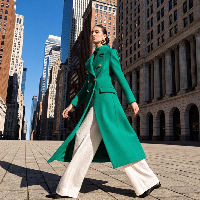 Full-length fashion editorial: model in a structured emerald wool coat and white trousers walking across a wide empty plaza, harsh midday sun, long shadow. — Recraft v4