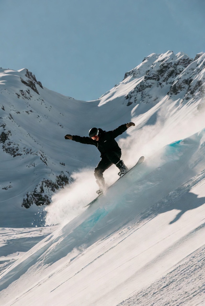 Snowboarder carving down a steep snowy slope with jagged alpine peaks in the background