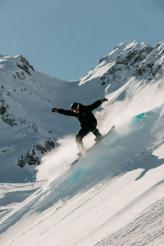 Snowboarder carving down a steep snowy slope with jagged alpine peaks in the background