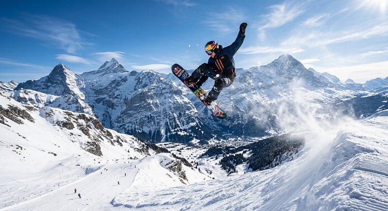 Snowboarder in mid-air performing a high jump on a snowy alpine slope with rugged mountains in the background.
