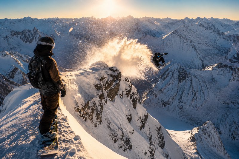 Snowboarder launches off a snow-covered cliff in an alpine mountain range at sunset, with a spectator watching.