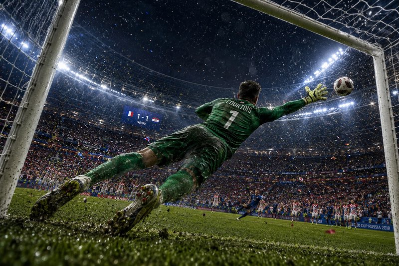 Goalkeeper in green dives to save the ball near the goal under bright stadium lights.