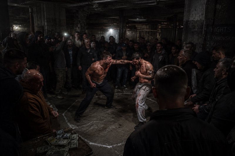 Two shirtless bare-knuckle fighters compete in an underground warehouse ring, as a crowd of spect...