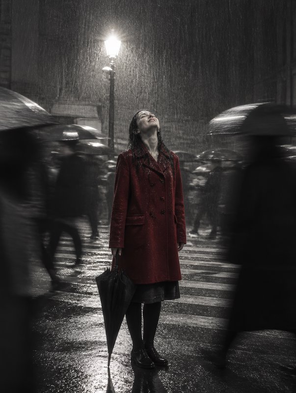 Woman in a red coat stands joyfully in heavy rain on a city crosswalk, holding a closed umbrella ...