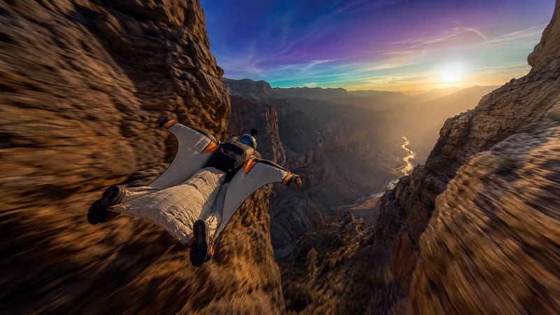 Wingsuit flyer glides at high speed between steep rocky canyon cliffs, motion blur emphasizing fast low flight at vibrant ...