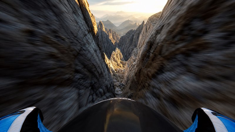 First-person POV of wingsuit flight speeding through a jagged rocky mountain canyon at golden hou...