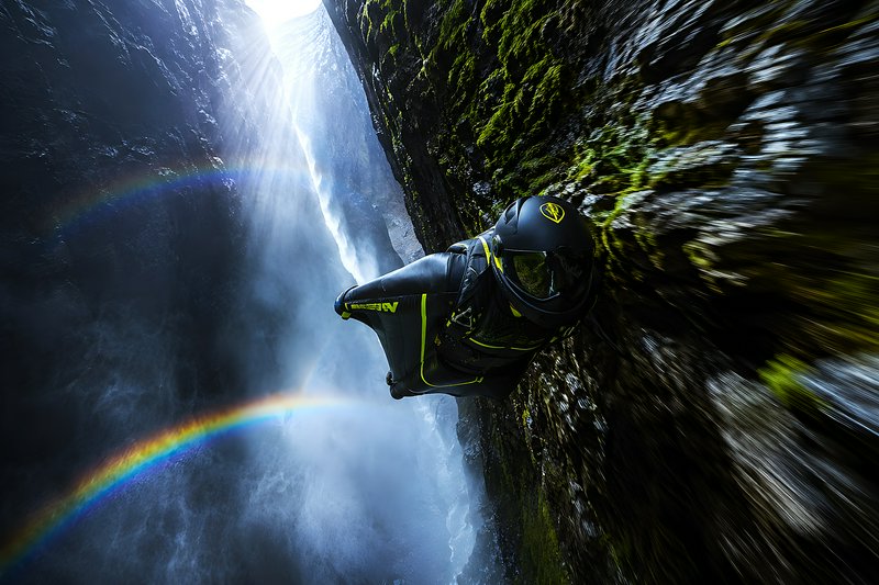 [Wingsuit flyer glides inches from a mossy canyon wall, with sunbeams, a waterfall, and double rainbows filling the narrow...