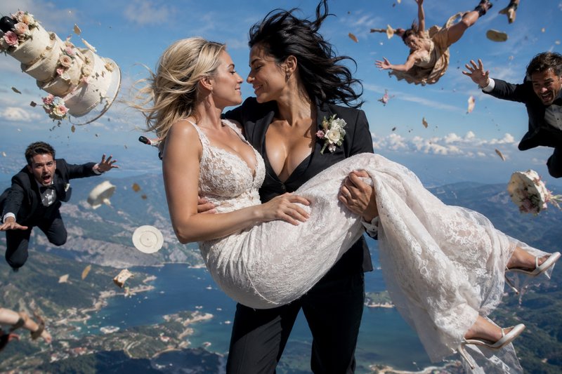 Queer wedding couple mid-skydive, one bride carrying her partner, with floating guests and a topp...