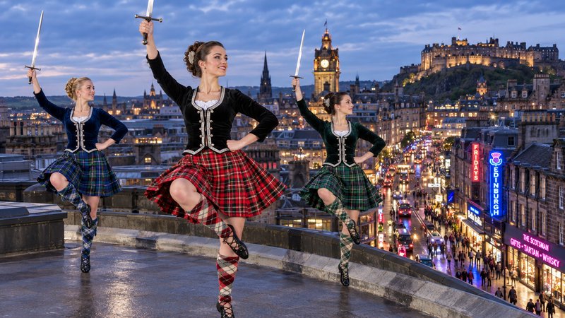 Three Scottish sword dancers pose on a rooftop overlooking Edinburgh's twilight city skyline