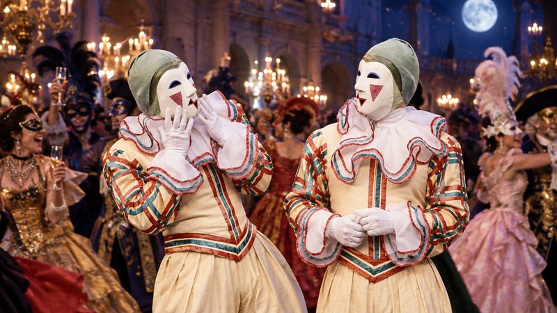 Matching masked Pierrot figures stand amid guests at a lavish Venetian masquerade ball