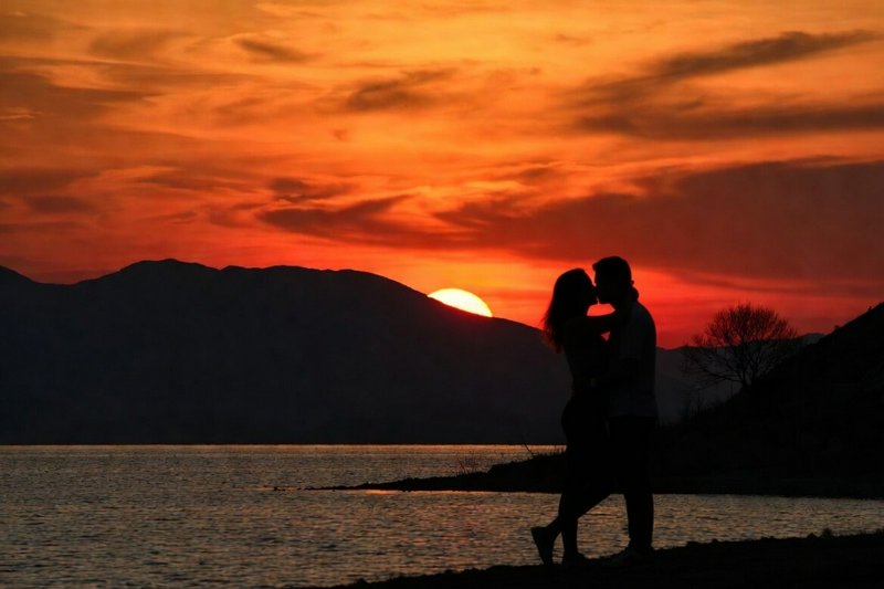 Silhouetted couple kissing at sunset along a lake with distant mountains.